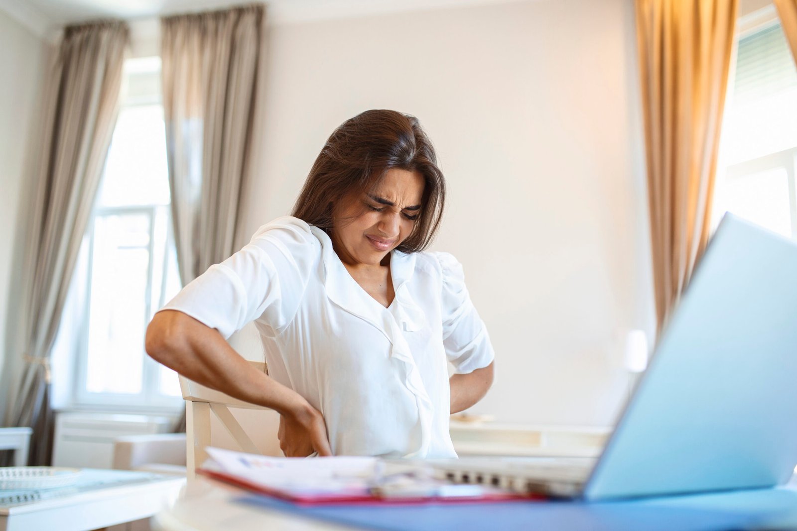 Portrait of young stressed woman sitting at home office desk in front of laptop, touching aching back with pained expression, suffering from backache after working on laptop