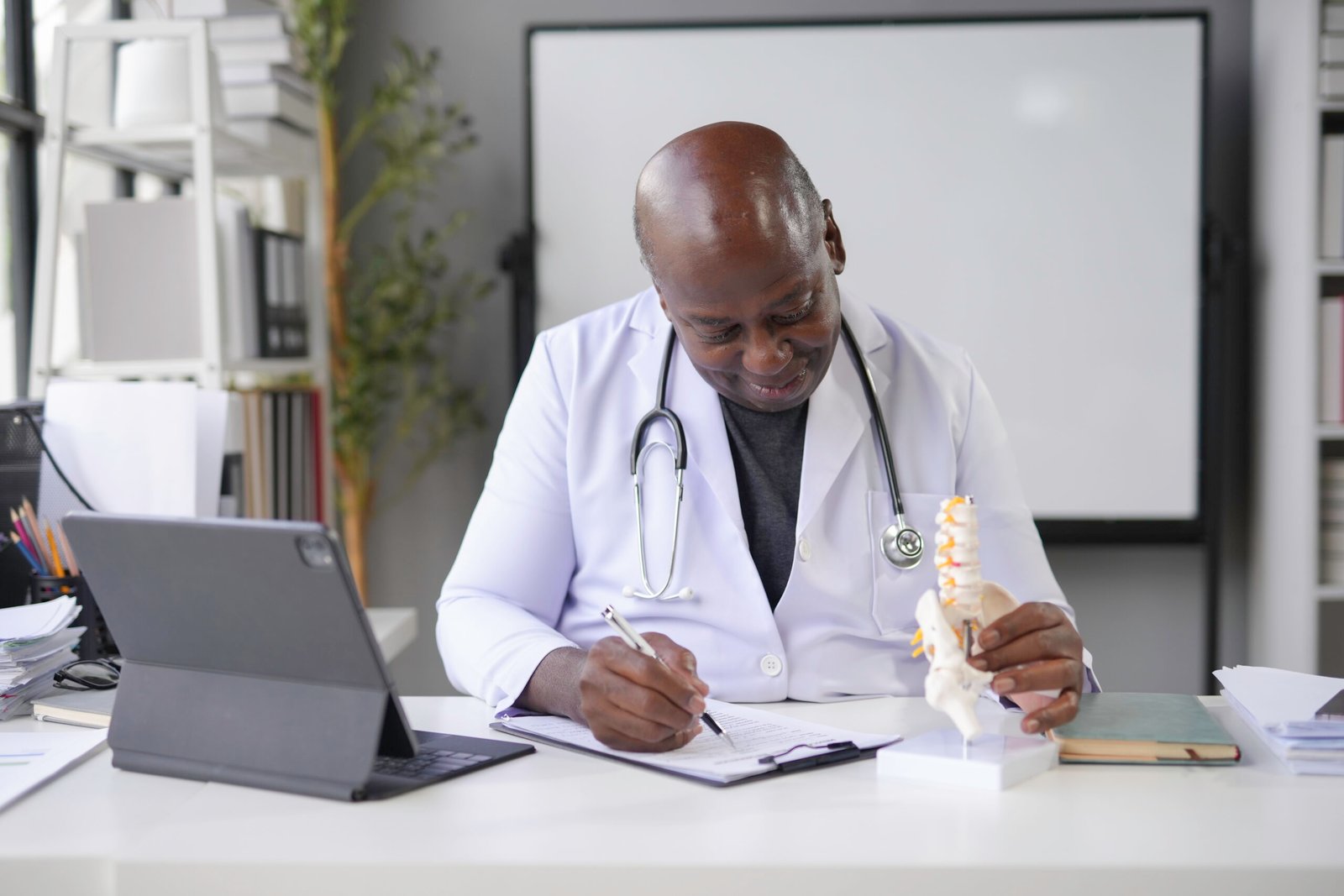 Doctor in a modern office setting, writing notes while examining a spine model. A laptop and medical books surround him, emphasizing a focus on healthcare and anatomy