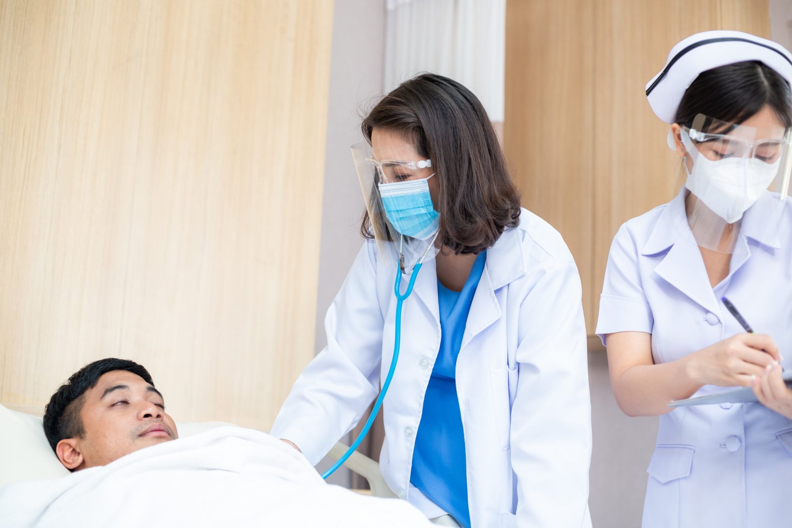 Asian female doctor wearing mask and face shield with nurse using stethoscope checking health of patient sleeping in bed at hospital ward
