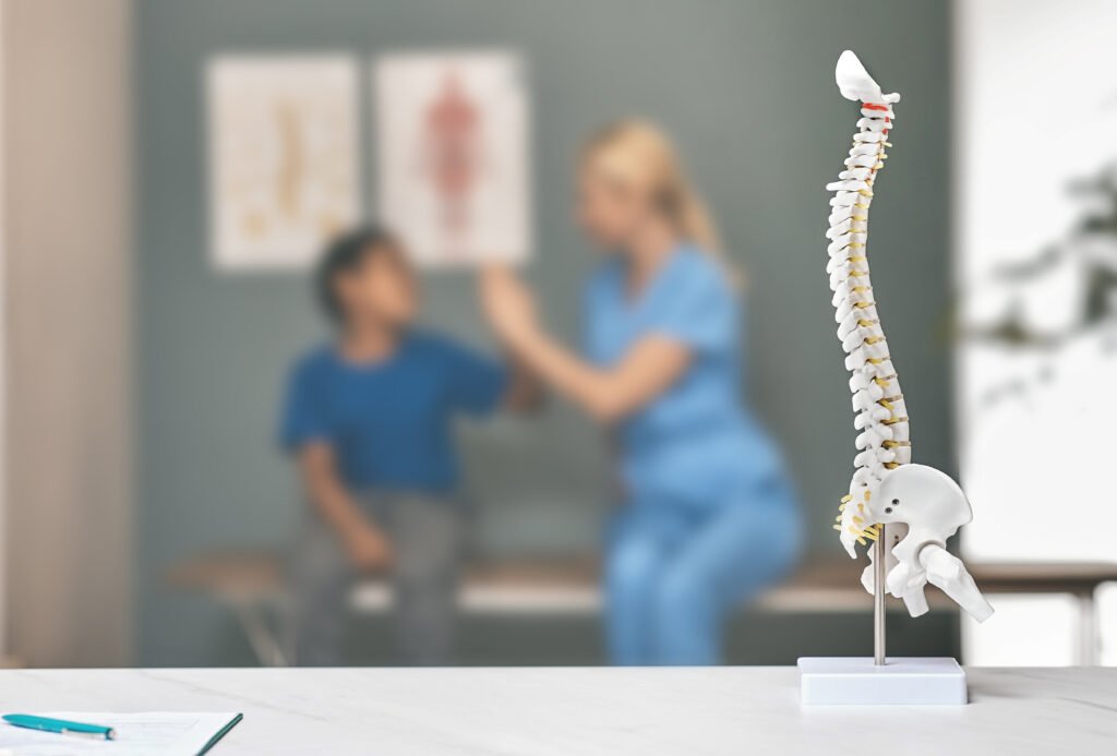 Anatomical model of the human spine on a clinic table with an experienced spine surgeon consulting a young patient in the background.