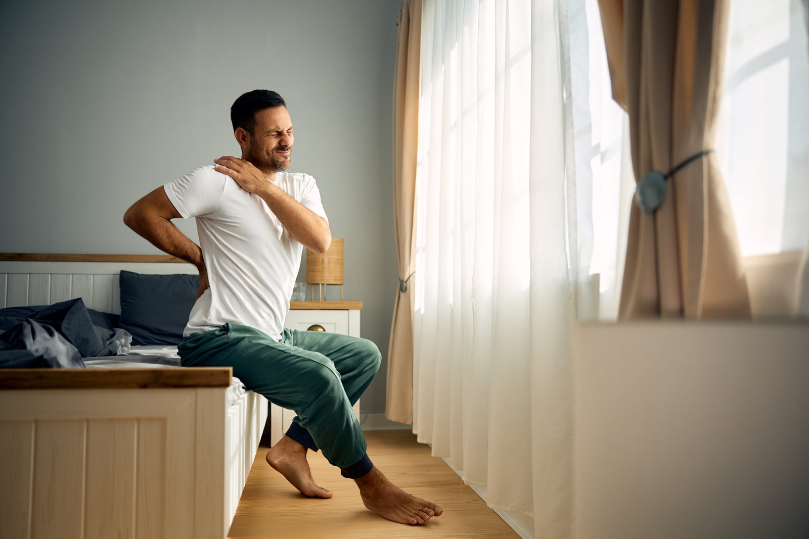 Mid adult man holding his back and shoulder in pain after waking up in the morning. Copy space.